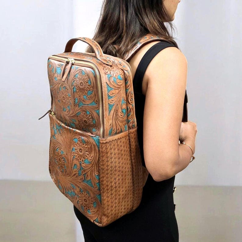 May include: A tan leather backpack featuring an intricate floral tooling pattern with turquoise accents. The bag includes a top handle, gold-tone zippers, and a side panel with a textured geometric pattern. It is worn with adjustable shoulder straps.