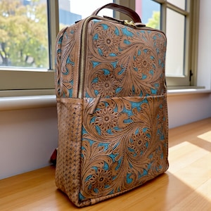 May include: A rectangular backpack featuring tan leather with intricate floral tooling and a vibrant turquoise blue textured background. The side panel displays a woven leather pattern, and the bag includes a top handle and gold-tone zippers.