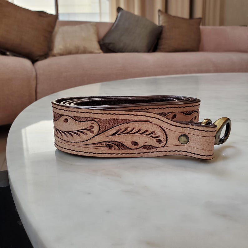 May include: A rolled-up, light brown leather belt with a floral embossed design. The belt has a dark brown interior and a brass buckle. The belt is sitting on a white marble surface.