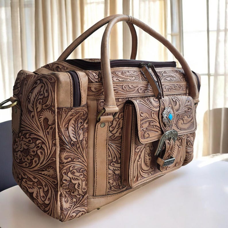 May include: A tan leather bag with intricate floral tooling. The bag features a top handle, a front pocket with a turquoise stone and silver buckle, and a side pocket. The bag has a zipper closure and is designed for travel.