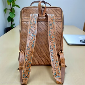 May include: A tan leather backpack featuring a woven texture pattern across the main body. The shoulder straps are decorated with intricate floral tooling and turquoise accents. The bag includes gold-tone zippers and a top carry handle.