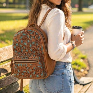Könnte beinhalten: Ein brauner Lederrucksack mit einem detaillierten floralen Prägemuster und türkisfarbenen Akzenten. Der Rucksack verfügt über eine vordere Reißverschlusstasche, zwei Schultergurte und silberfarbene Metallreißverschlüsse. Das Muster bedeckt die Vorderseite.