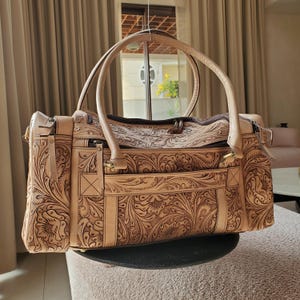 May include: A tan leather tote bag with intricate floral tooling. The bag features two rounded handles, a zippered top, and a front pocket. The detailed carving creates a textured surface, and the bag appears to be new.