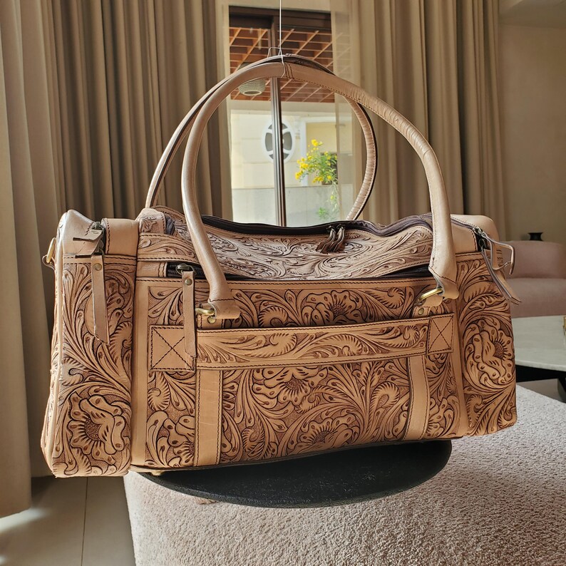 May include: A tan leather tote bag with intricate floral tooling. The bag features two rounded handles, a zippered top, and a front pocket. The detailed carving creates a textured surface, and the bag appears to be new.