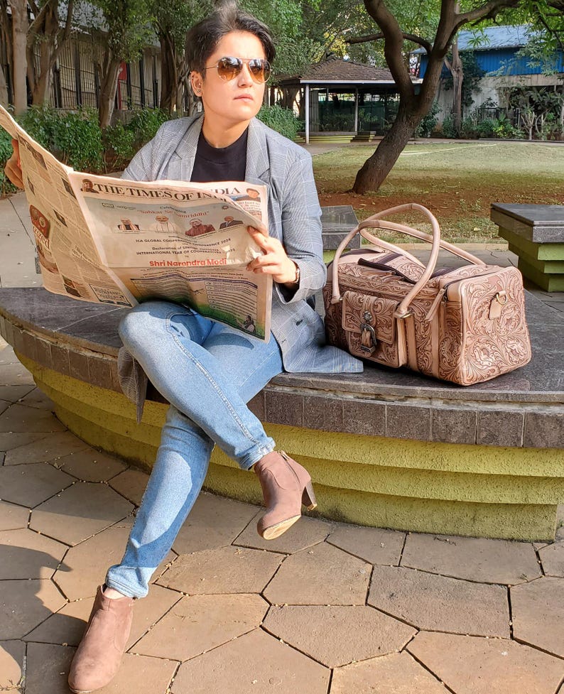 May include: A light brown leather handbag with a floral design and a metal clasp sits on a stone bench. The Times of India newspaper is held open. The person is wearing a gray blazer, blue jeans, and brown suede boots.