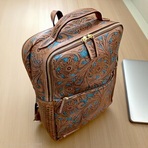 May include: A tan leather backpack featuring intricate floral tooling and embossed patterns with bright turquoise accents. The bag includes a top handle, gold-tone metal zippers, and a woven leather side panel detail on the exterior structure.