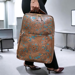 May include: A rectangular leather backpack featuring an intricate floral tooling pattern in tan brown with a vibrant turquoise blue background. The bag includes a top handle, gold-tone metal zippers, and a large front pocket with matching floral details.