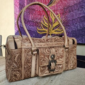 May include: A beige leather handbag with intricate floral and scrollwork carvings. The bag features two top handles, a front pocket with a decorative buckle, and a zipper closure. The bag is a light tan color.