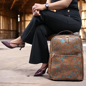 May include: A rectangular backpack featuring a tan leather exterior with an intricate floral tooling pattern and vibrant turquoise accents. The bag includes a top handle, a front zippered pocket, and gold-toned metal zippers along the main compartment.
