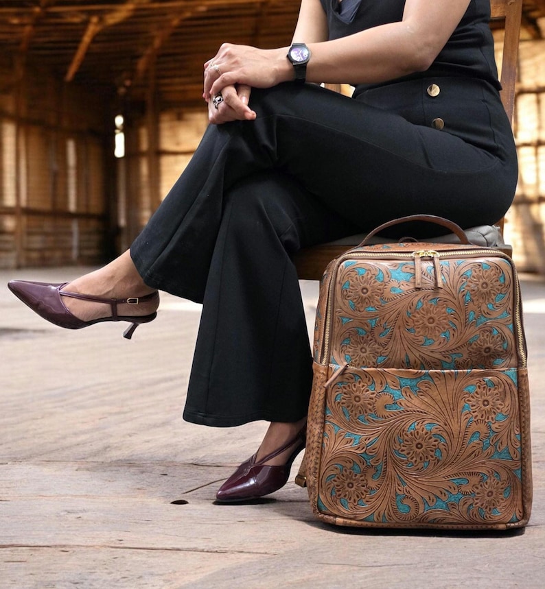 May include: A rectangular backpack featuring a tan leather exterior with an intricate floral tooling pattern and vibrant turquoise accents. The bag includes a top handle, a front zippered pocket, and gold-toned metal zippers along the main compartment.