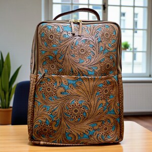 May include: A rectangular backpack featuring a tan leather exterior with an intricate embossed floral pattern. The recessed areas of the design reveal a bright turquoise blue background. The bag includes gold-tone metal zippers and a top handle.