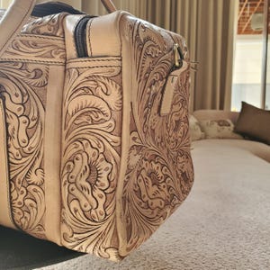 May include: A tan leather duffel bag with intricate floral and scrollwork carving. The bag features a zippered top, side pocket, and a handle. The leather has a natural, aged appearance, with a detailed embossed design.