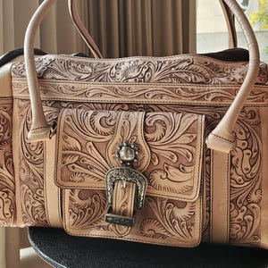 May include: A light tan leather handbag with intricate floral and scroll tooling. The bag features a front flap with a decorative silver buckle and a matching silver embellishment. The bag has two handles and a shoulder strap.