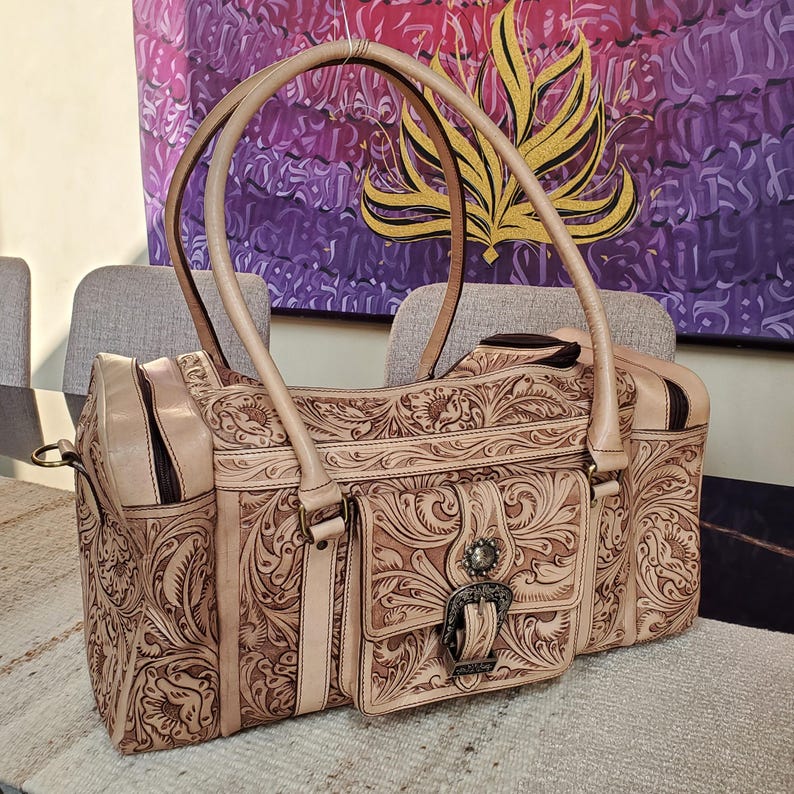 May include: A tan leather handbag with intricate floral and scroll tooling. The bag features a front pocket with a decorative buckle, double handles, and a zippered top. The bag is sitting on a table.
