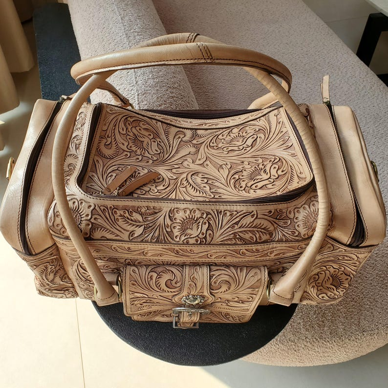 May include: Tan leather handbag with intricate floral and leaf carvings. The bag features a top handle, a front pocket with a buckle closure, and a zipper closure. The bag is a light brown color.