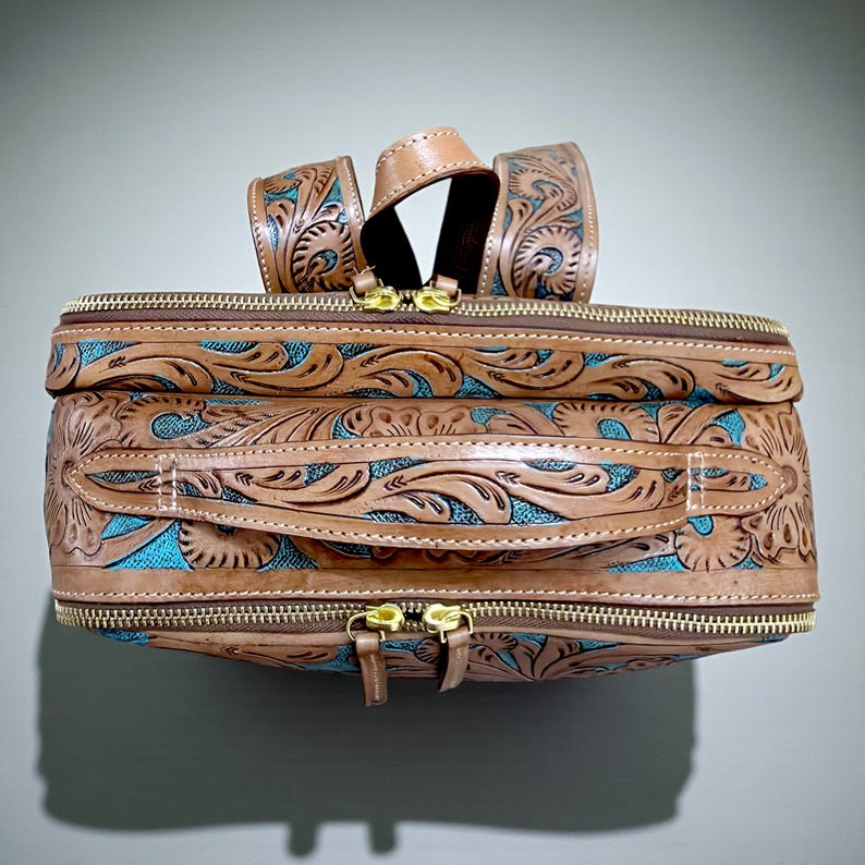 May include: A rectangular backpack featuring tan leather with intricate floral tooling and a vibrant turquoise fabric inlay. The bag includes a top handle, gold-tone metal zippers, and adjustable shoulder straps with matching leather and turquoise detailing.