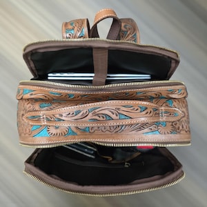 May include: A brown leather backpack featuring intricate floral tooling with turquoise accents. The bag has multiple zippered compartments, gold-tone hardware, and a top handle. The interior is lined with dark fabric and includes a laptop sleeve.
