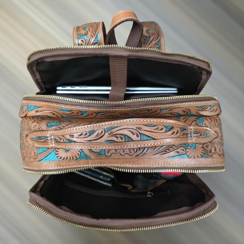 May include: A brown leather backpack featuring intricate floral tooling with turquoise accents. The bag has multiple zippered compartments, gold-tone hardware, and a top handle. The interior is lined with dark fabric and includes a laptop sleeve.