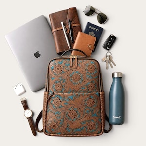 May include: A brown leather backpack with an intricate floral pattern and turquoise accents, surrounded by a laptop, a leather notebook, a passport, sunglasses, a car key, a set of keys, a water bottle, a wristwatch, and wireless earbuds.