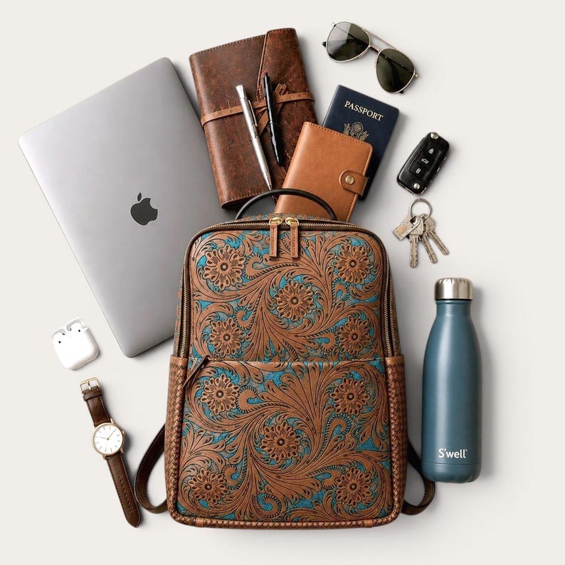May include: A brown leather backpack with an intricate floral pattern and turquoise accents, surrounded by a laptop, a leather notebook, a passport, sunglasses, a car key, a set of keys, a water bottle, a wristwatch, and wireless earbuds.