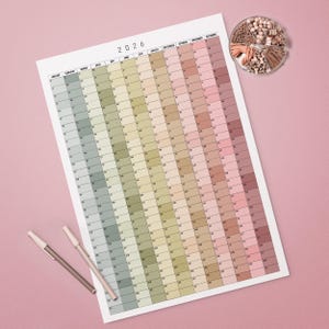 May include: A large 2026 year calendar with a gradient of pastel colours. The calendar is on a white background with two pens and a small container of decorative items. The calendar is a useful tool for planning and organisation.