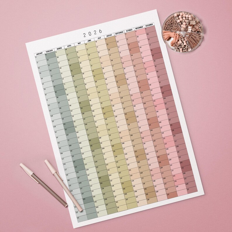 May include: A large 2026 year calendar with a gradient of pastel colours. The calendar is on a white background with two pens and a small container of decorative items. The calendar is a useful tool for planning and organisation.