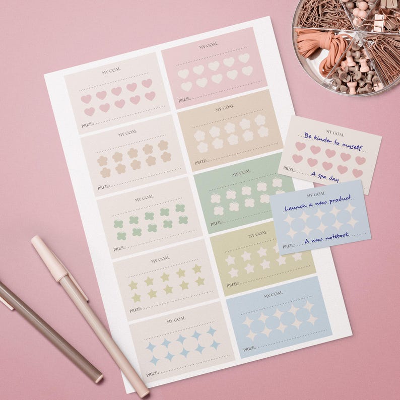 May include: A sheet of goal-setting cards with pastel-coloured squares, each featuring a different shape like hearts, stars, and flowers. Two pens and a container of office supplies are also visible. The cards have the words "My Goal" and "Prize".