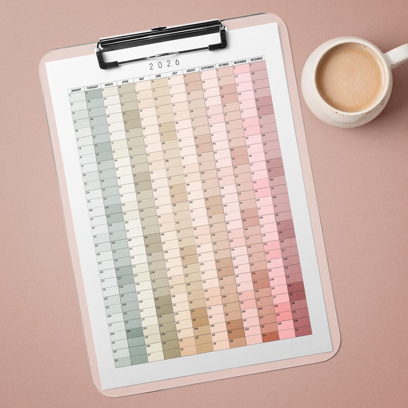 May include: A 2026 year planner on a clipboard, with a pastel colour gradient for each month. The planner is clipped to a light pink clipboard with a silver clip. A white mug of coffee sits to the side.