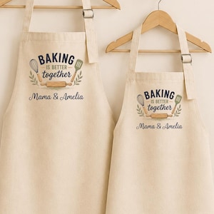 May include: Two beige aprons of different sizes hanging on wooden hangers. Each apron features a graphic with a whisk, spatula, and rolling pin, along with the text "BAKING IS BETTER together" and the personalised names "Mama & Amelia" in dark ink.