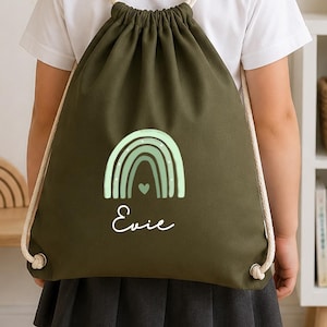 May include: An olive green drawstring backpack featuring a light green rainbow graphic with a small heart at the centre and the name Evie printed in white script lettering below the rainbow. The bag has thick cream-coloured rope shoulder straps.