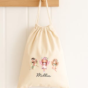 May include: A cream-coloured drawstring bag with three watercolour fairy illustrations, each wearing floral crowns and wings. The name "Millie" is written in cursive below. The bag hangs from a wooden peg.