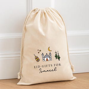 Peut inclure: Un sac cadeau beige à cordon de serrage avec une illustration colorée d'une mosquée, d'une lanterne, d'étoiles et d'un croissant de lune. Le texte "EID GIFTS FOR Ismaeel" est imprimé en dessous.