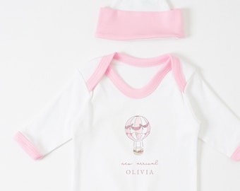 Puede incluir: Mono blanco para bebé con ribetes rosas y un gráfico de globo aerostático. El mono dice "new arrival" y "OLIVIA". Viene con un gorro blanco con un borde rosa.