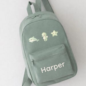 May include: A pale green backpack with a front zipped pocket and adjustable straps. The backpack has embroidered sea creature designs: a dolphin, seahorse, and starfish. The name "Harper" is printed in white on the front pocket.