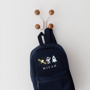 May include: A navy blue backpack with a space theme design. It features a cartoon astronaut, a rocket ship, and a satellite. The backpack has a zippered pocket and the name "MICAH" is printed on the front.