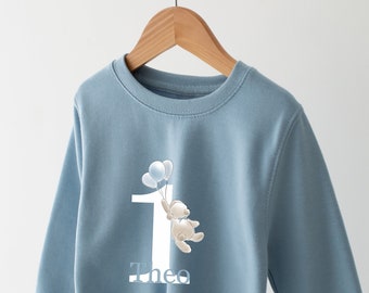 May include: Light blue sweatshirt with a white number one, a teddy bear holding balloons, and the name "Theo" printed on the front.