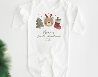 May include: White baby onesie with a brown bear, a green Christmas tree, and a red stocking. The onesie has the text "Nora's first christmas 2024" printed on it.