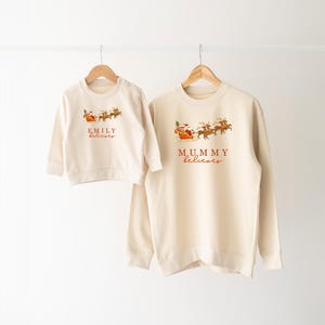 May include: Two cream-colored Christmas sweatshirts with Santa and reindeer graphics. The larger sweatshirt says "MUMMY believes" and the smaller sweatshirt says "EMILY believes".