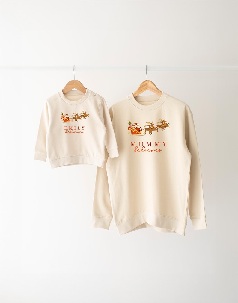May include: Two cream-colored Christmas sweatshirts with Santa and reindeer graphics. The larger sweatshirt says "MUMMY believes" and the smaller sweatshirt says "EMILY believes".