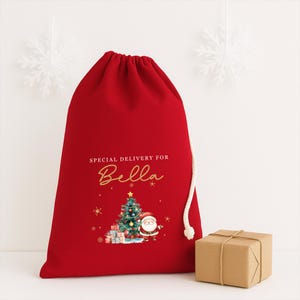 May include: A red Christmas gift bag with a drawstring closure. The bag features the text "SPECIAL DELIVERY FOR Bella" in gold script, along with a Christmas tree, Santa Claus, and gift boxes illustration. A small, wrapped gift box sits beside the bag.