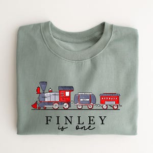 May include: A light green t-shirt featuring a cartoon train design and the text "FINLEY is one".