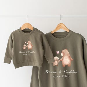 May include: Two olive green sweatshirts with a bear and a smaller bear holding hands with hearts above them. The larger sweatshirt has the text "Mama & Freddie since 2023".