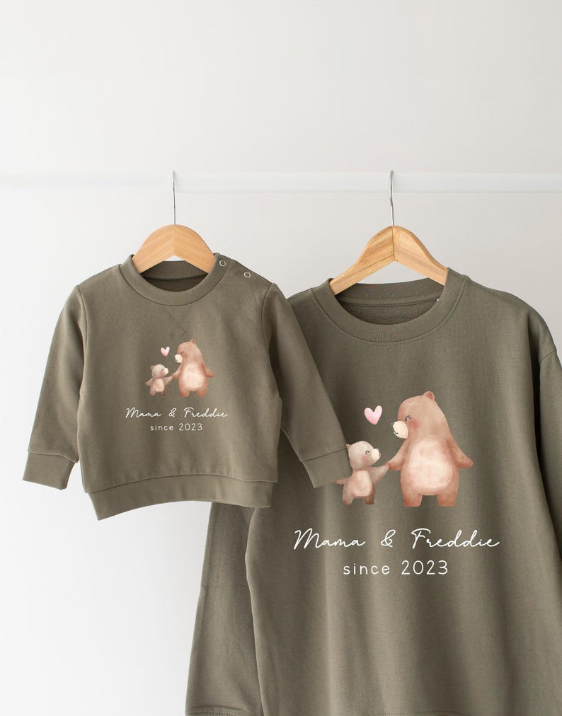 May include: Two olive green sweatshirts with a bear and a smaller bear holding hands with hearts above them. The larger sweatshirt has the text "Mama & Freddie since 2023".