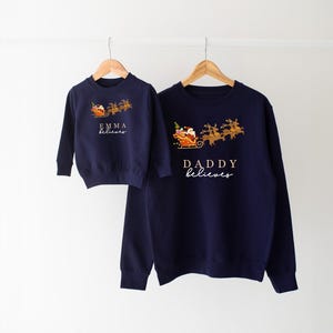 May include: Two navy blue Christmas sweaters with a Santa Claus and reindeer design. The smaller sweater says "Emma believes" and the larger sweater says "Daddy believes".