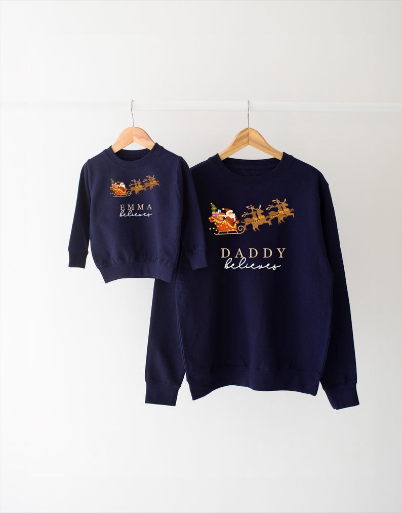 May include: Two navy blue Christmas sweaters with a Santa Claus and reindeer design. The smaller sweater says "Emma believes" and the larger sweater says "Daddy believes".