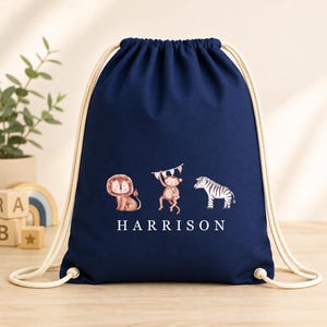May include: A navy blue drawstring backpack featuring a printed graphic of a lion, a monkey holding a flag, and a zebra. The name HARRISON is printed in white capital letters below the animals. The bag has thick, cream-coloured rope straps.