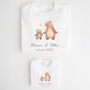 May include: Two white t-shirts with a watercolor illustration of two bears holding hands. The larger bear is brown and the smaller bear is light brown. The text "Mama & Ellie since 2022" is printed on both shirts.