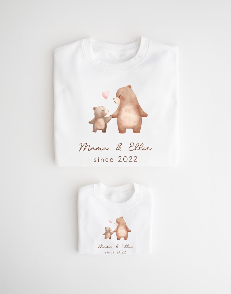 May include: Two white t-shirts with a watercolor illustration of two bears holding hands. The larger bear is brown and the smaller bear is light brown. The text "Mama & Ellie since 2022" is printed on both shirts.