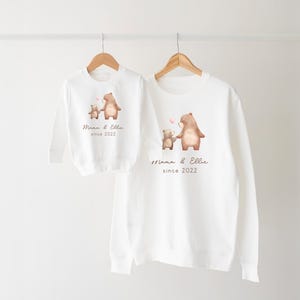 May include: White long-sleeve sweatshirts with a brown bear and a smaller brown bear holding a pink heart. The text on the sweatshirts reads "Mama & Ellie since 2022".