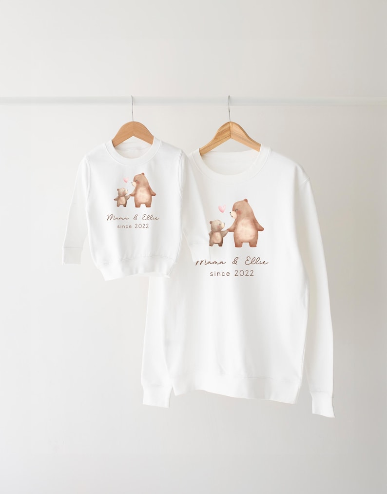 May include: White long-sleeve sweatshirts with a brown bear and a smaller brown bear holding a pink heart. The text on the sweatshirts reads "Mama & Ellie since 2022".
